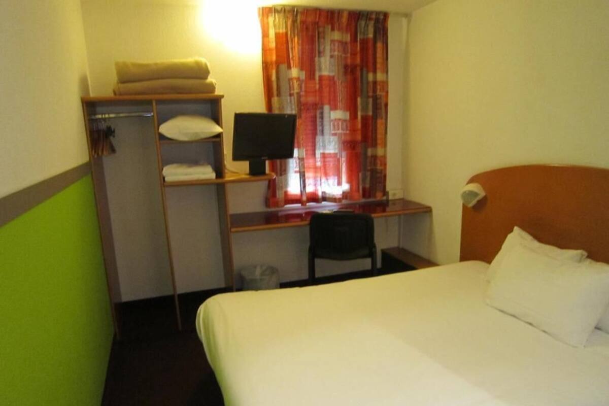 Une chambre d’hôtel avec un lit, un bureau avec un ordinateur, une chaise et une étagère avec des serviettes.