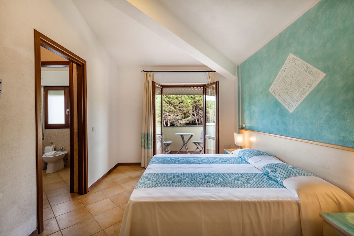 A hotel room with a bed, a small table, a balcony with a view of trees, and a bathroom visible through an open door.