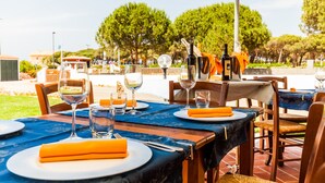 A dining area with tables set for a meal, outdoor seating, and a view of trees and buildings in the background. - Petri Marini Hotel & Resort (Aglientu)