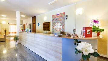 A hotel reception area with a reception desk, a large decorative wall piece, a potted plant, and a lamp.
