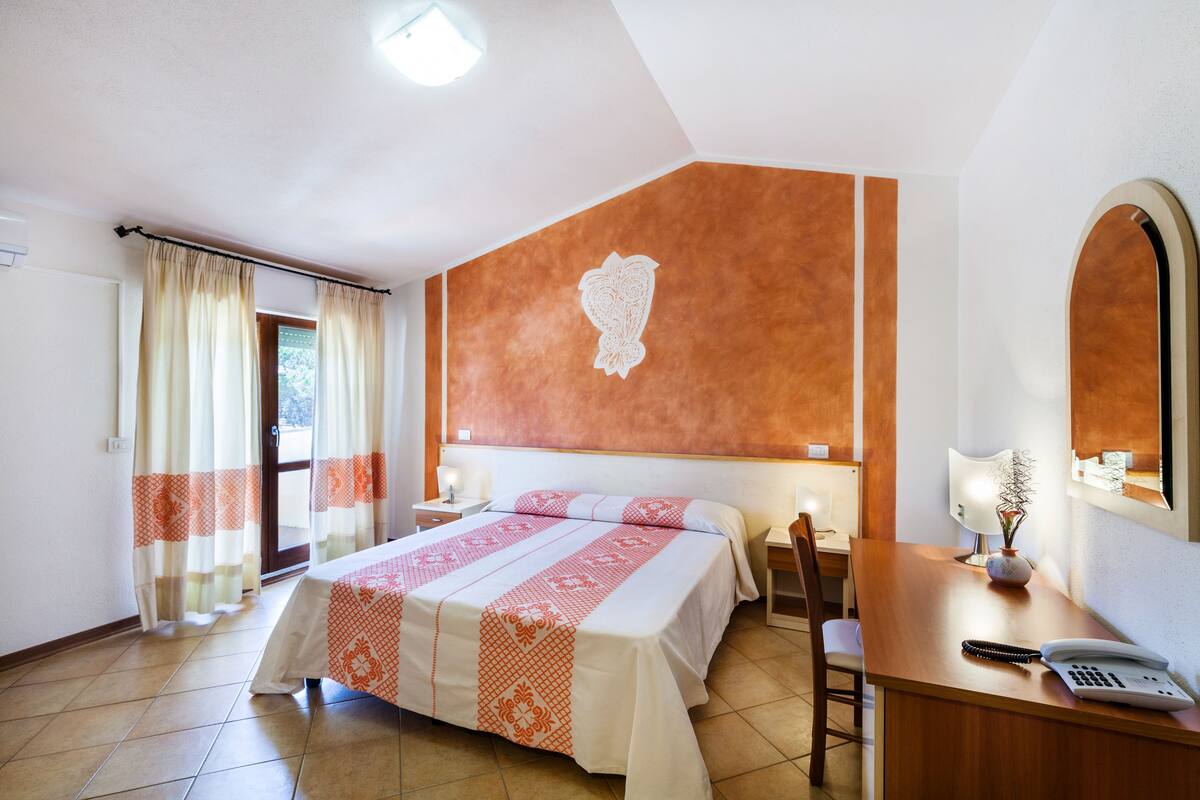 A hotel room with a bed, a desk, a chair, a lamp, and a mirror.