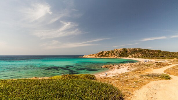 A coastal landscape with clear turquoise waters, a sandy beach, and a dirt path leading to a hill with sparse vegetation. - Petri Marini Hotel & Resort (Aglientu)