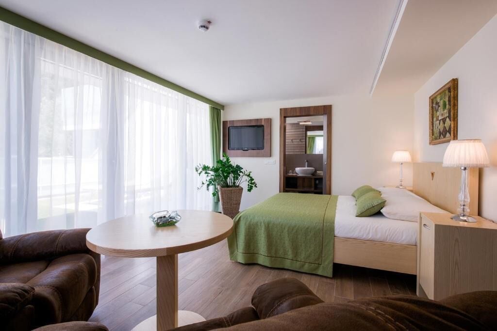 Ein Hotelzimmer mit Bett, Wandfernseher, Nachttisch mit Lampe, Fenster mit leichtem Vorhang und einer Zimmerpflanze.