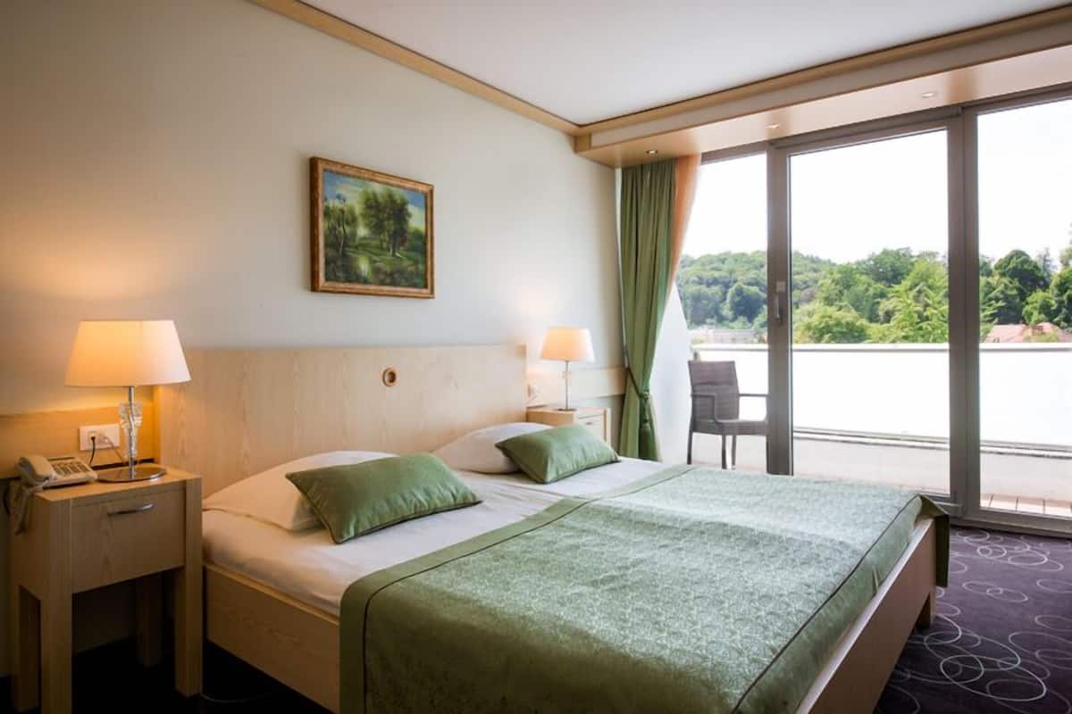 Une chambre d’hôtel avec un grand lit, deux lampes de chevet, un tableau au mur et une vue sur une étendue d’eau, visible à travers de grandes fenêtres.