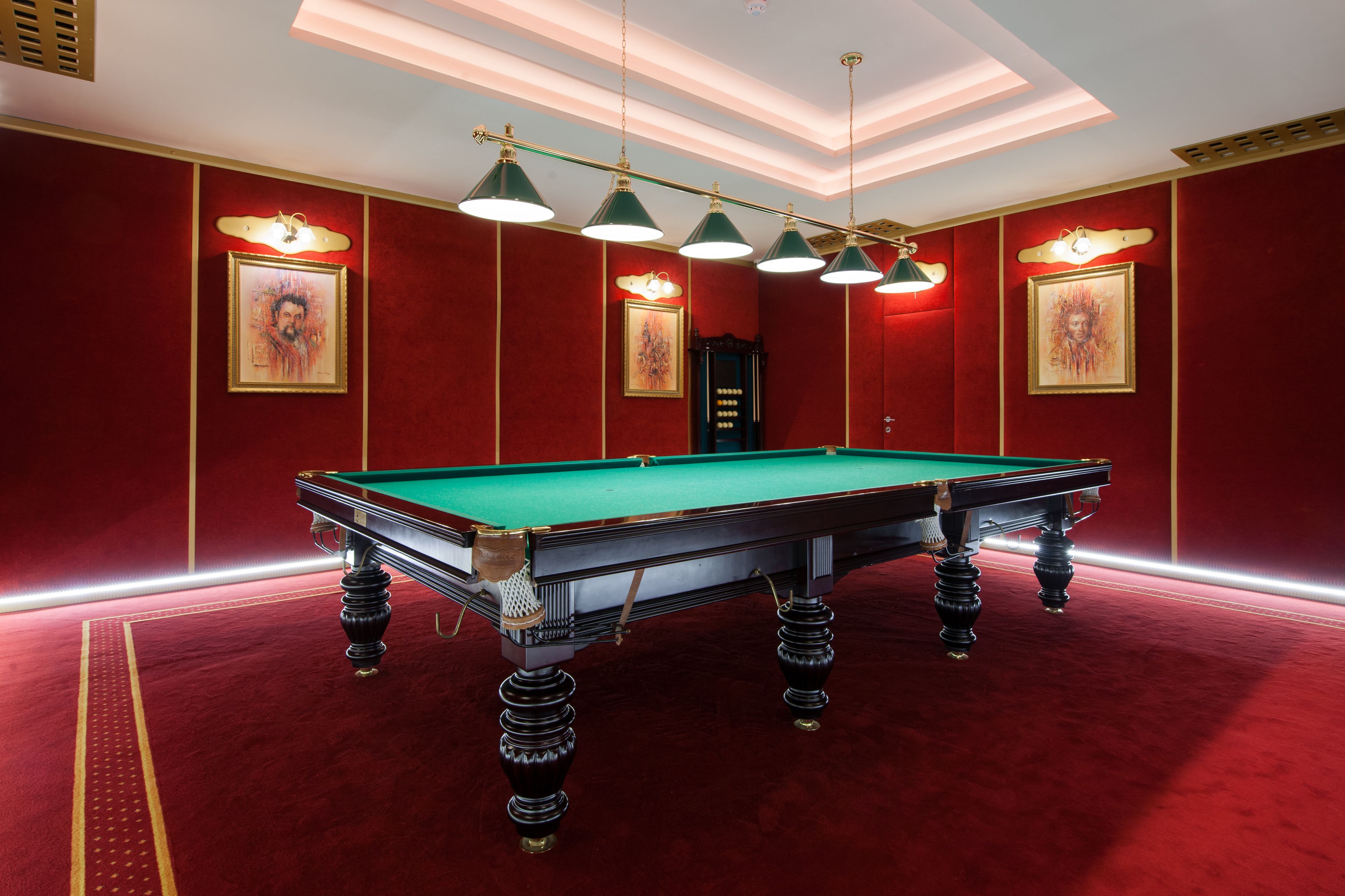 Une table de billard recouverte de feutre vert, entourée d’un tapis rouge et de murs rouges, avec des luminaires au plafond et des œuvres d’art encadrées.