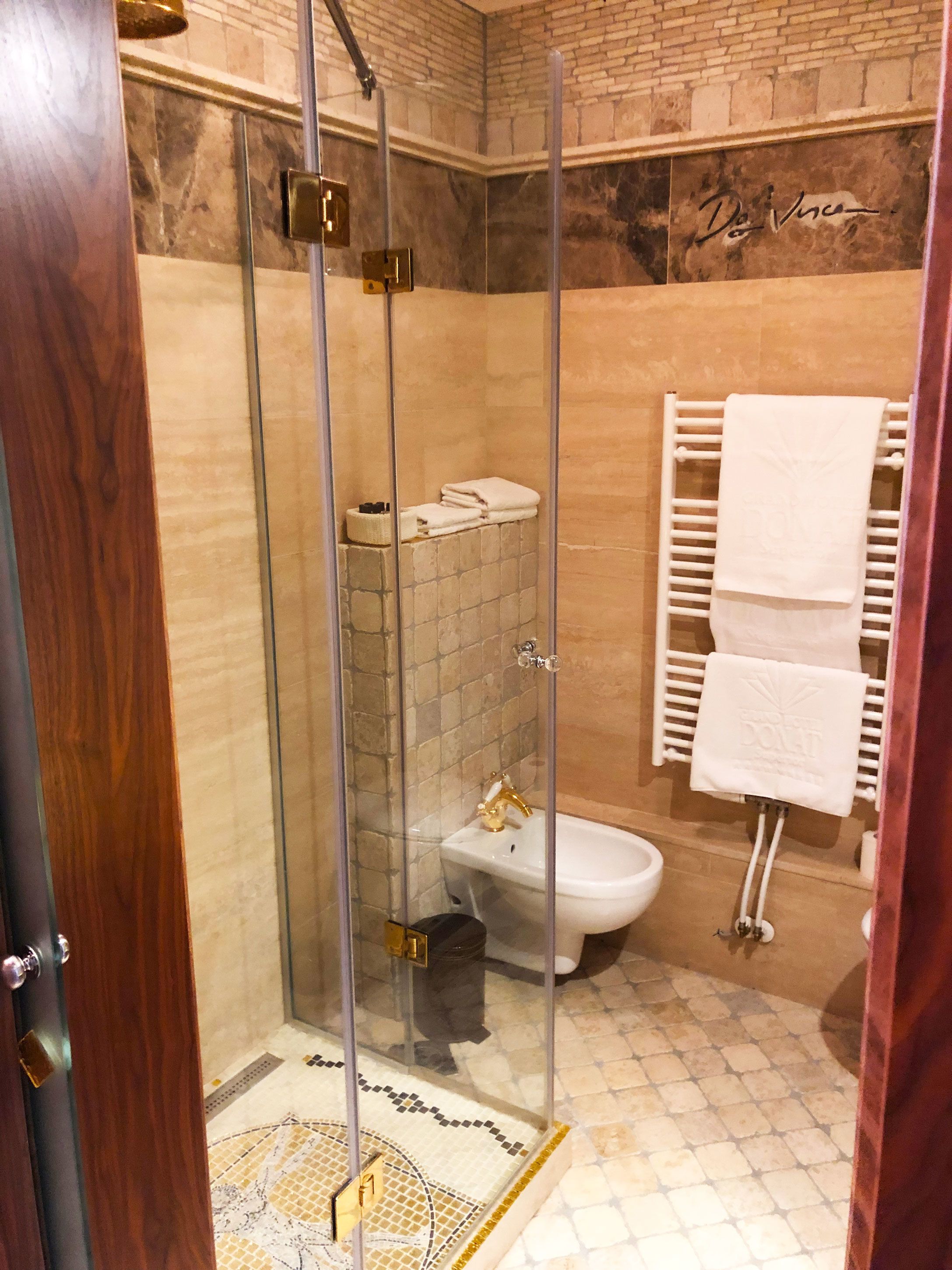 Et brusebad med glasvægge, toilet og vask, flisegulv og et håndklædestativ med håndklæder.