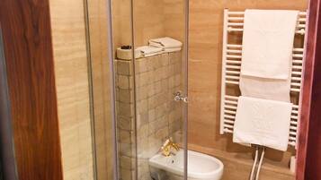 Et brusebad med glasvægge, toilet og vask, flisegulv og et håndklædestativ med håndklæder.