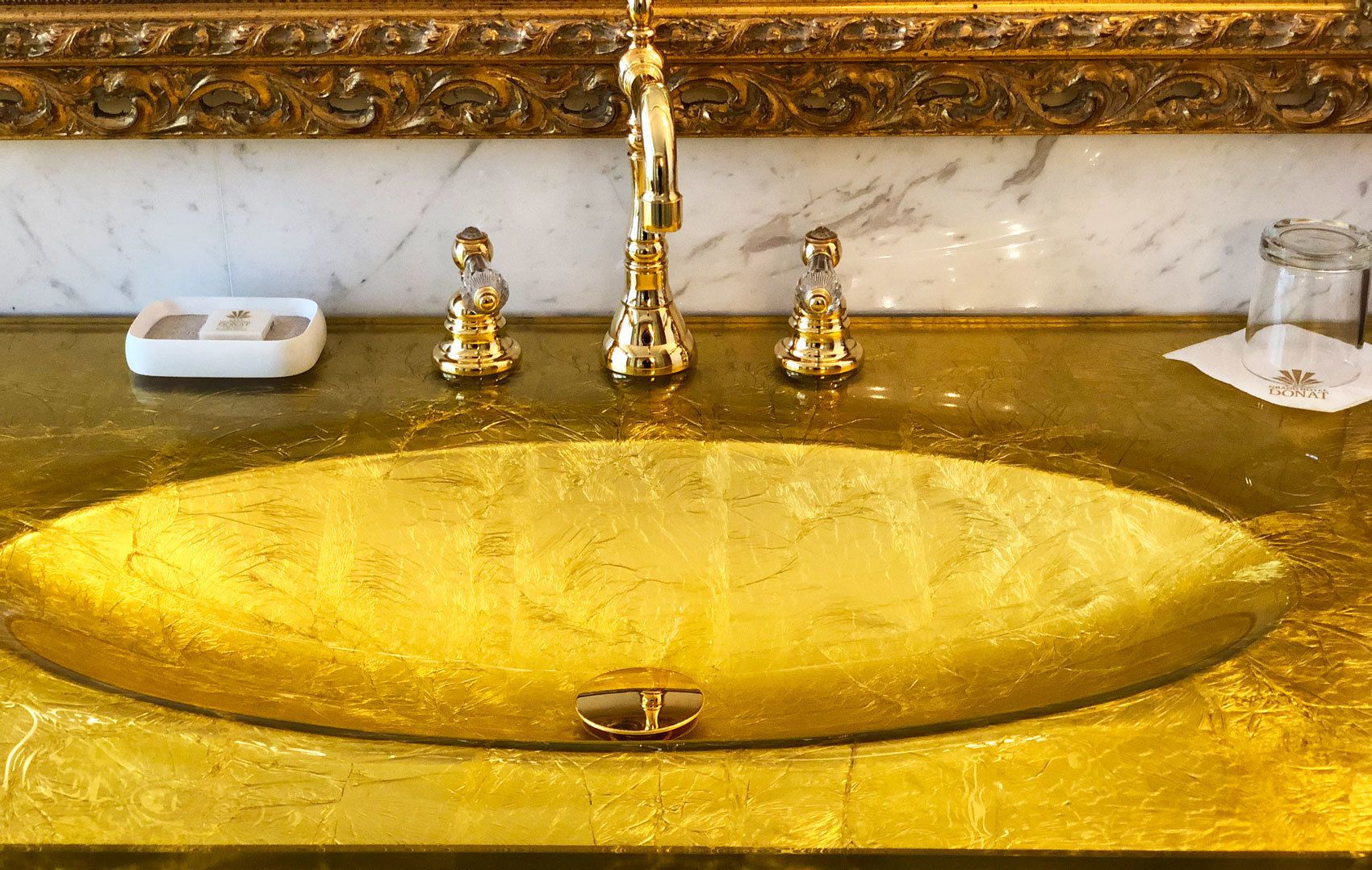 A gold-colored sink with three faucets, a soap dish, and a glass container with a label.