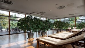 Een moderne, overdekte ontspanningsruimte met ligbedden, grote plantenpotten en grote ramen met uitzicht op de omgeving.