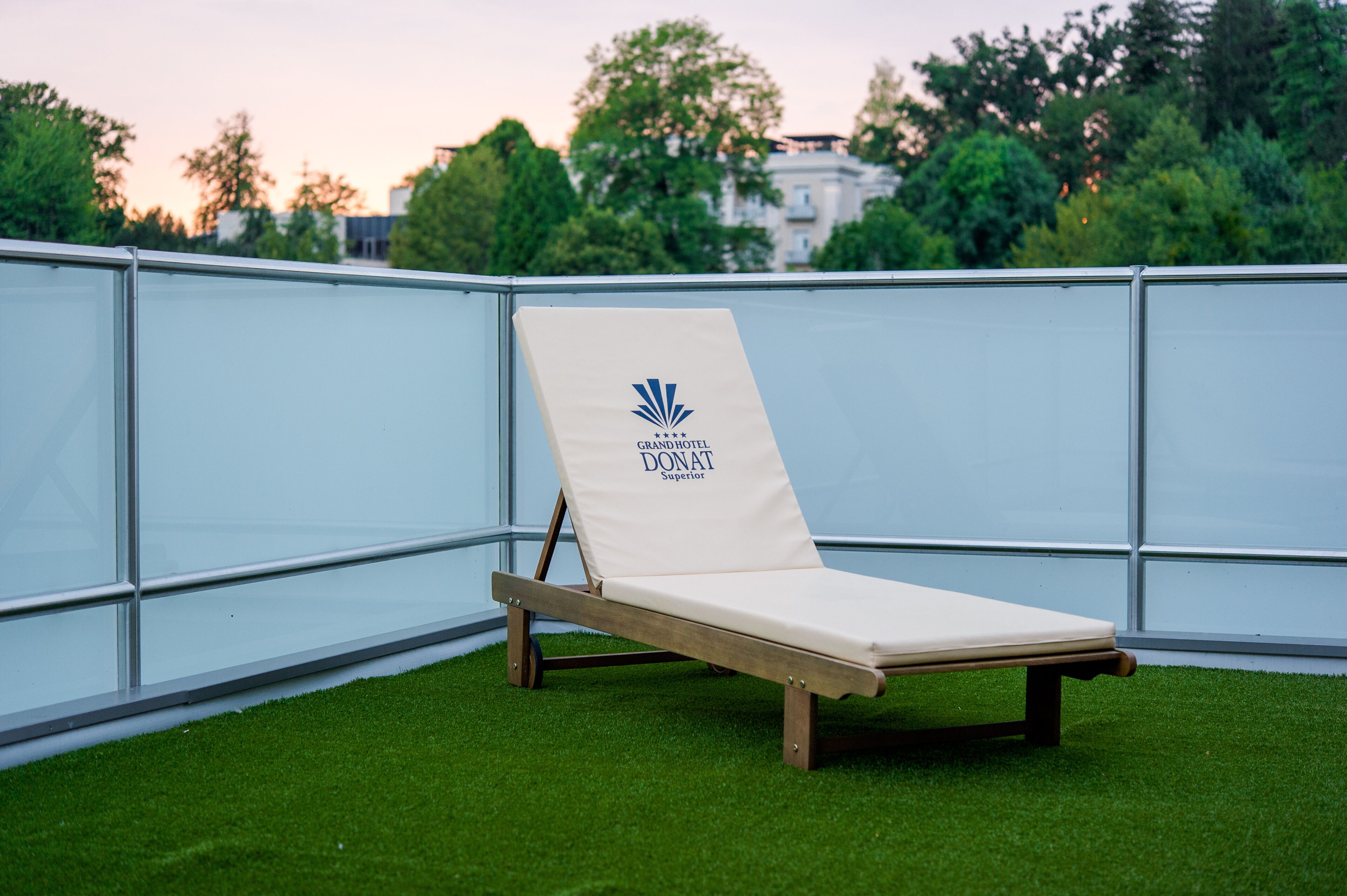 A lounge chair with a logo and text, positioned on artificial grass with a glass barrier and trees in the background.