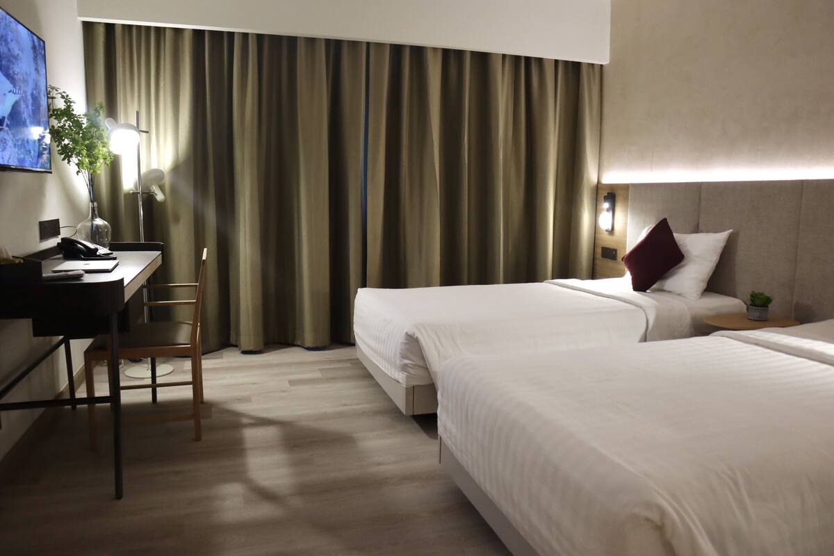A hotel room with two beds, a desk, a chair, a TV, and a window with curtains.