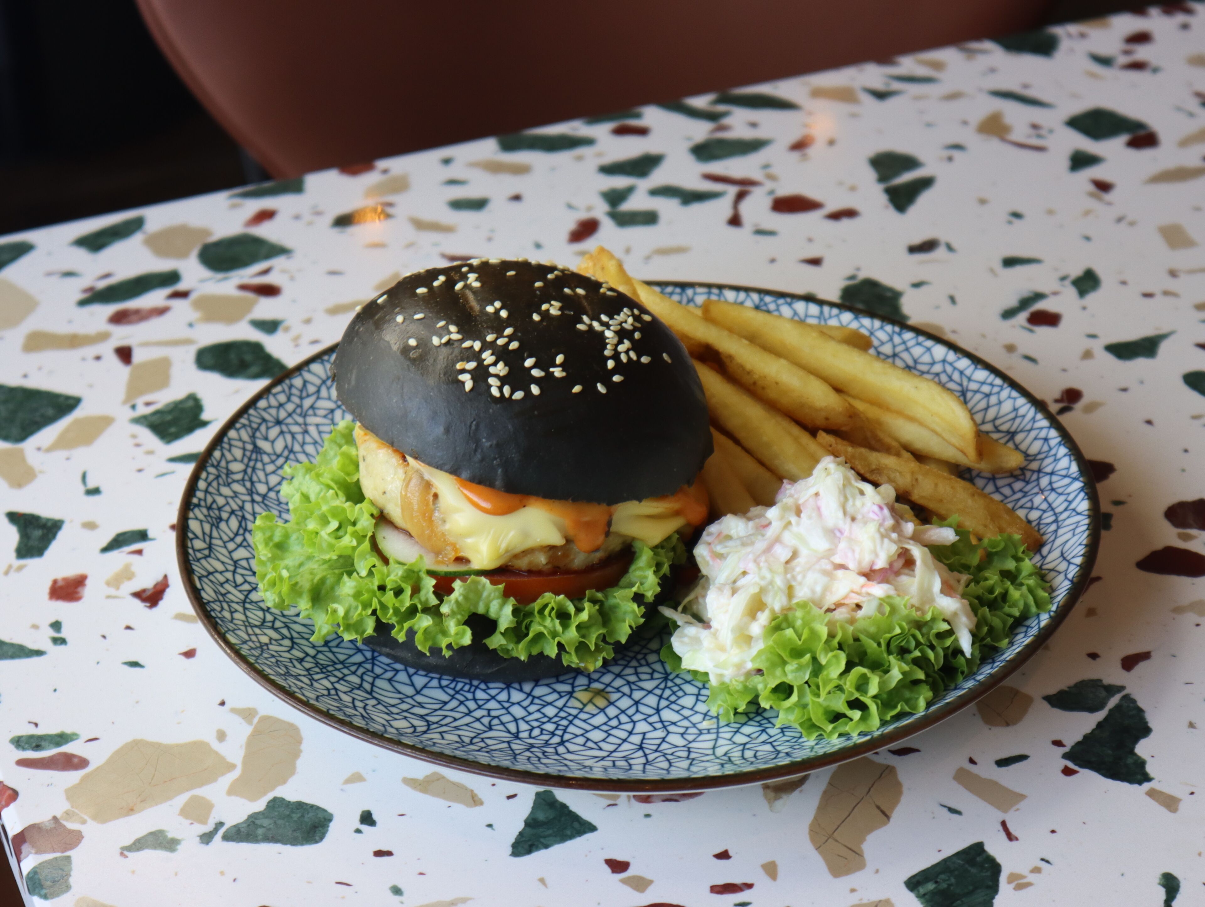 A plate with a burger, fries, and coleslaw on a speckled table.