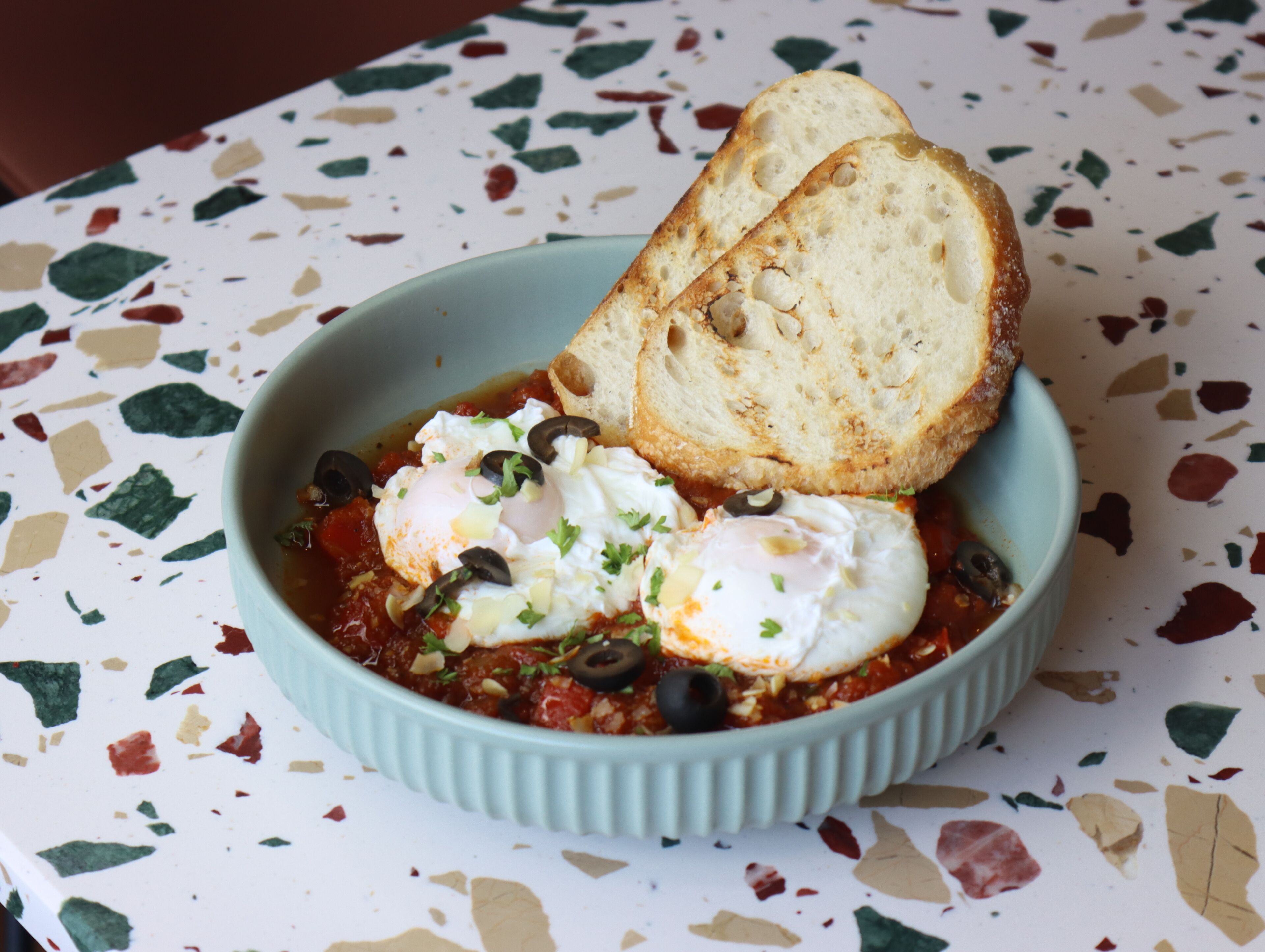 A bowl of food with eggs, olives, and bread on a speckled table.