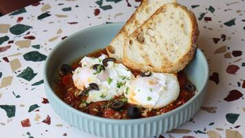 A bowl of food with eggs, olives, and bread on a speckled table.