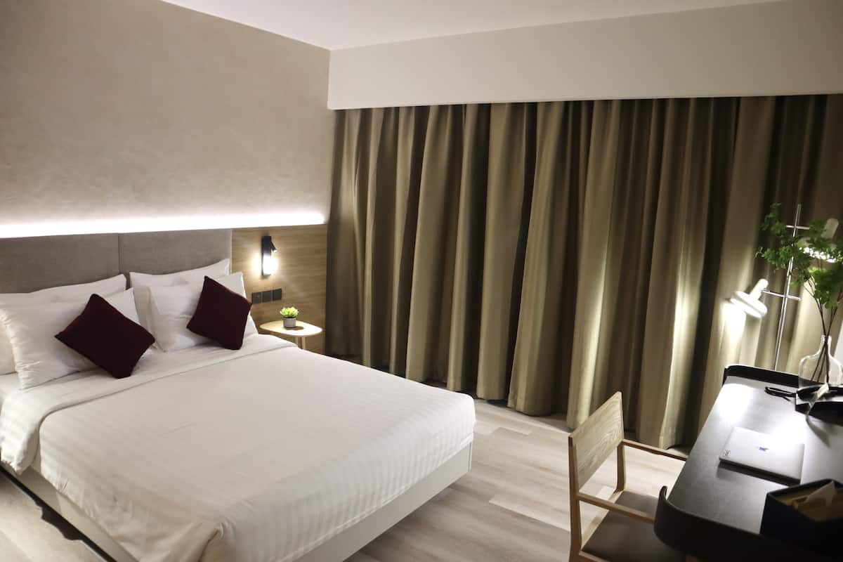 A hotel room with a large bed, a desk, and a chair. There is a lamp on the desk and a plant on the side.