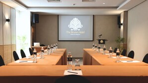 A conference room with two long tables, each with water bottles, glasses, and notepads. A projector screen displays the hotel's logo.