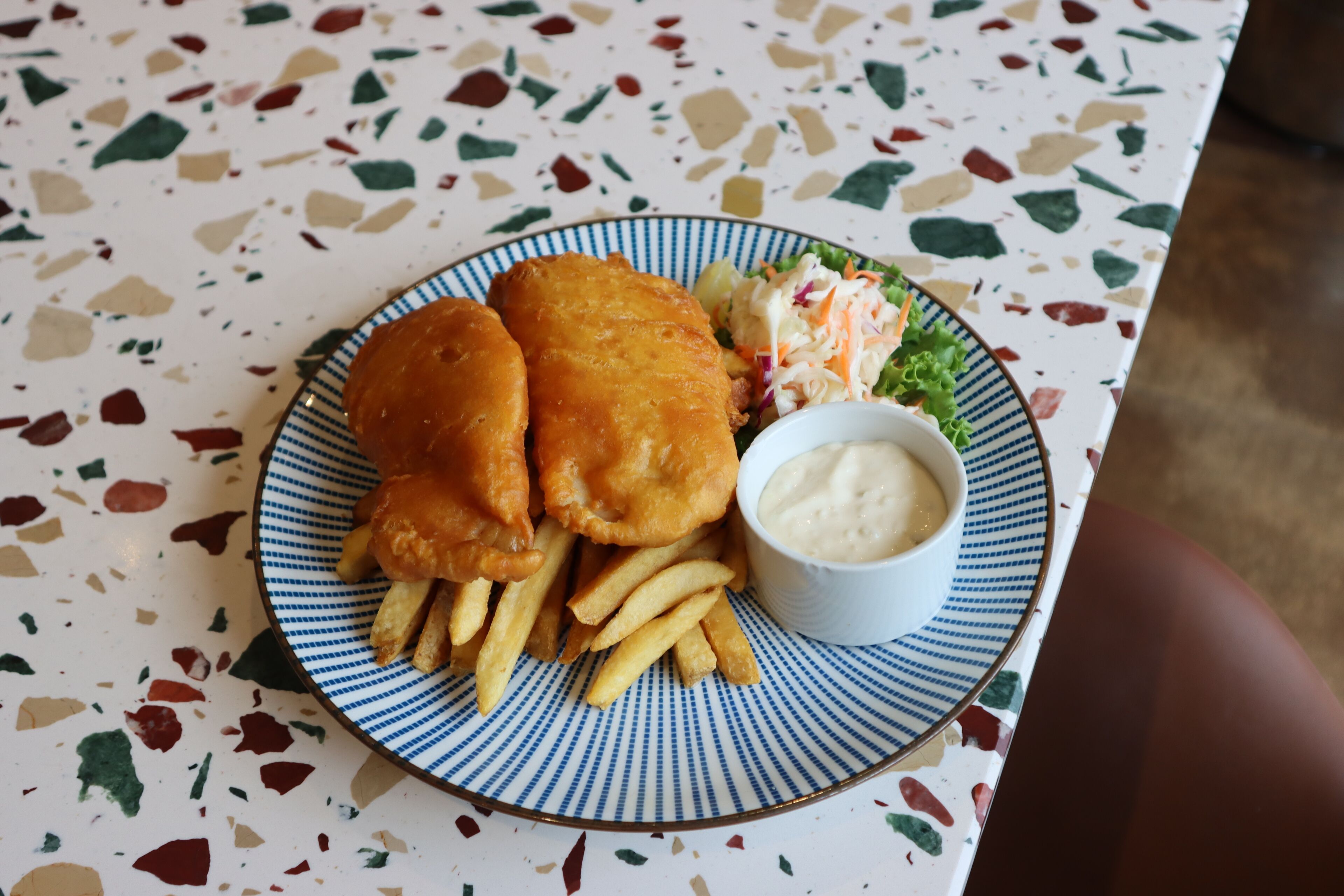 A plate with fish and chips, fries, and a small bowl of sauce on a speckled table.