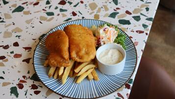 A plate with fish and chips, fries, and a small bowl of sauce on a speckled table.