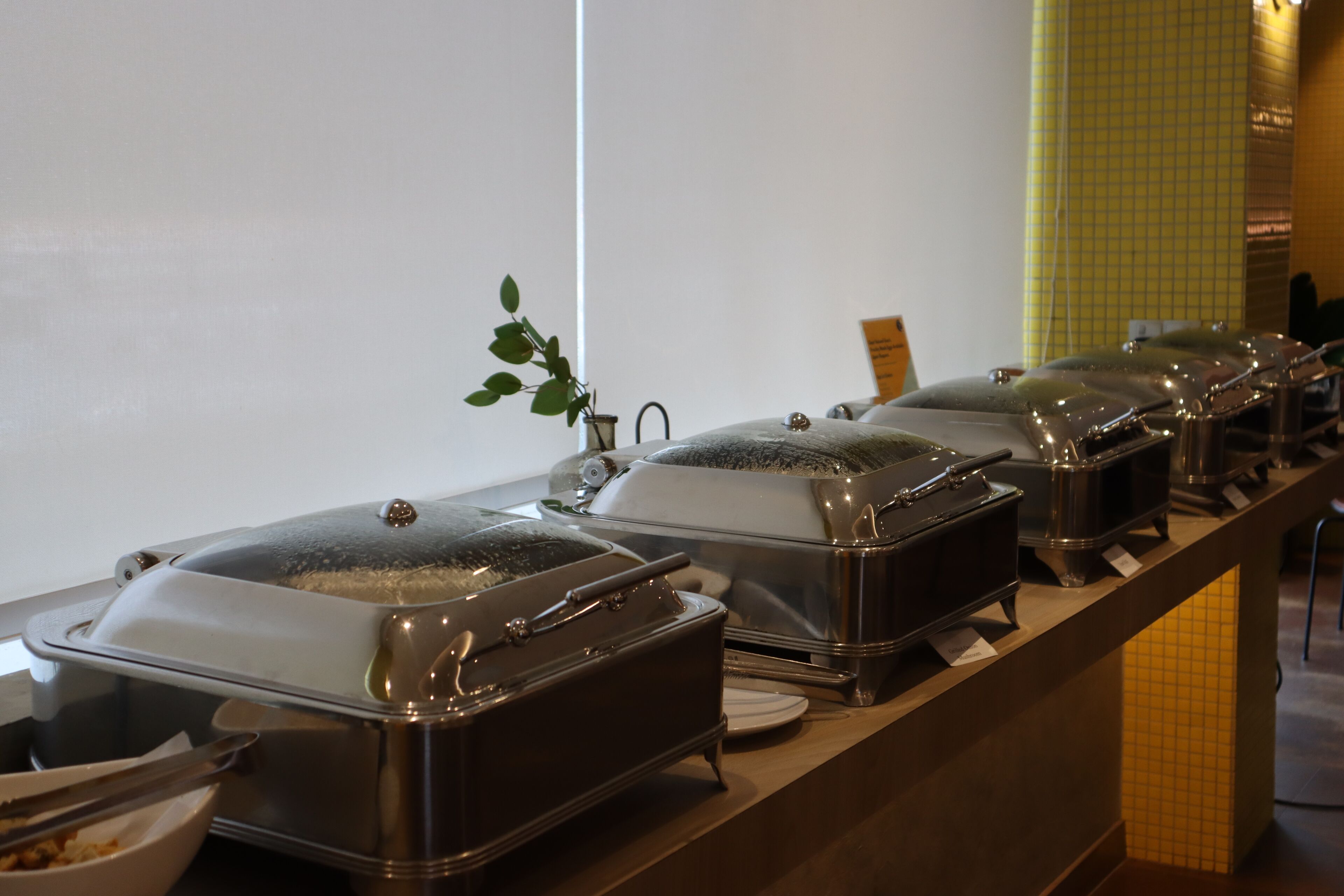A buffet setup with covered food dispensers, a potted plant, and a tiled wall.