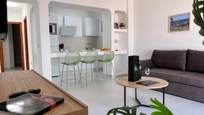 Een moderne woonkamer met een grijze bank, een salontafel met een boek en een glas, en een keukenhoek met witte kasten en groene stoelen.