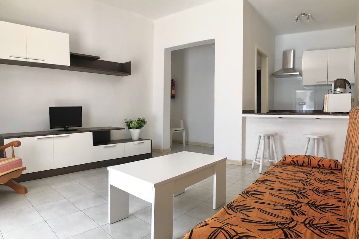 Una cocina moderna con armarios blancos, encimera negra y estufa integrada. Hay una mesa de comedor blanca y un sofá con funda estampada.