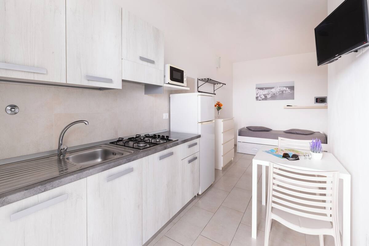 A modern kitchen with white cabinets, a stainless steel sink, a microwave, and a refrigerator. There is a small table with a chair and a TV mounted on the wall.