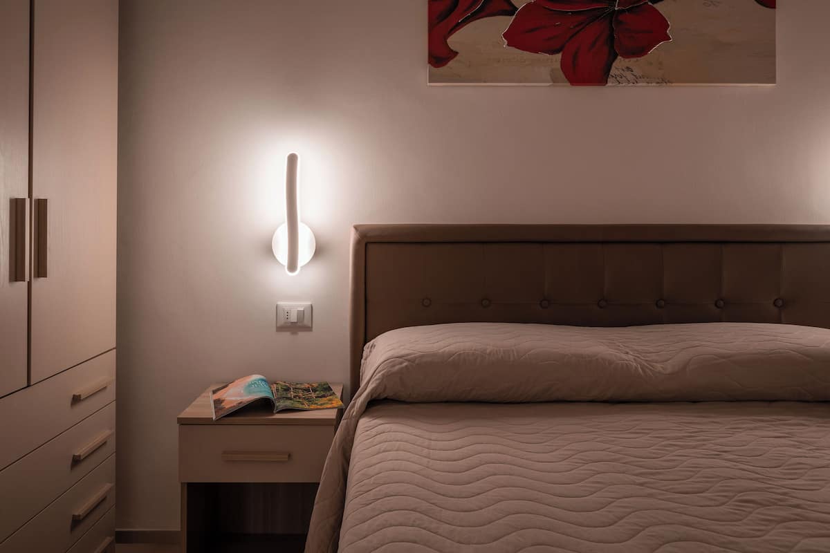 A bedroom with a bed, a nightstand with a lamp, and a wall-mounted light.