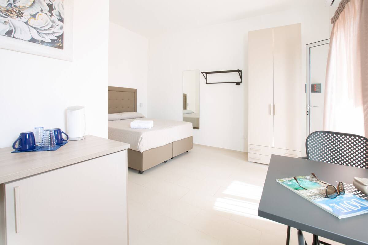 A hotel room with a bed, a desk, and a chair. There is a mirror on the wall and a window with curtains.