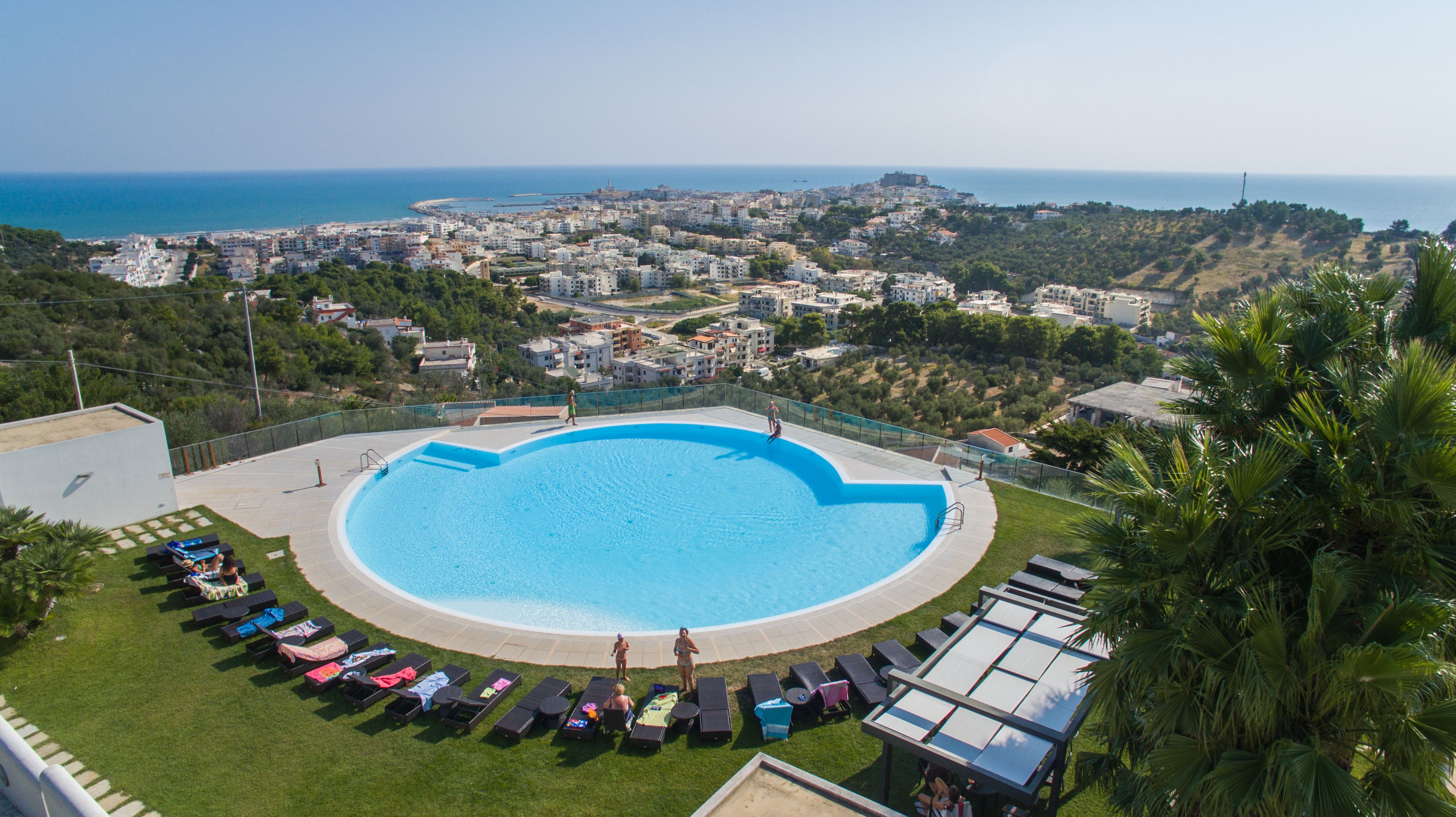 A large circular pool with a clear blue center, surrounded by lounge chairs and umbrellas, overlooking a coastal cityscape with buildings and greenery.