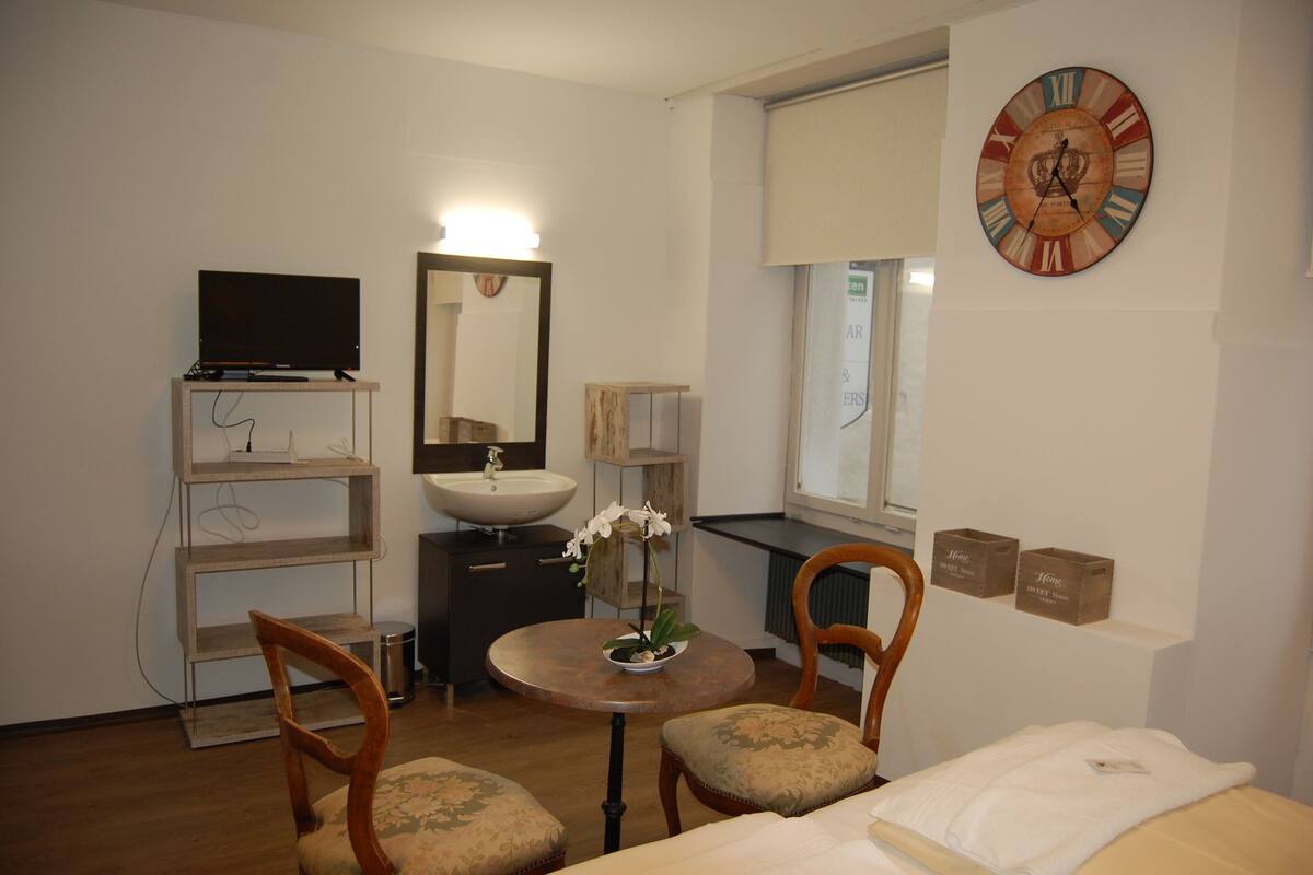 A hotel room with a round table, two chairs, a bed, a TV, a sink, a mirror, a clock, and two storage boxes labeled "Home Sweet Home".