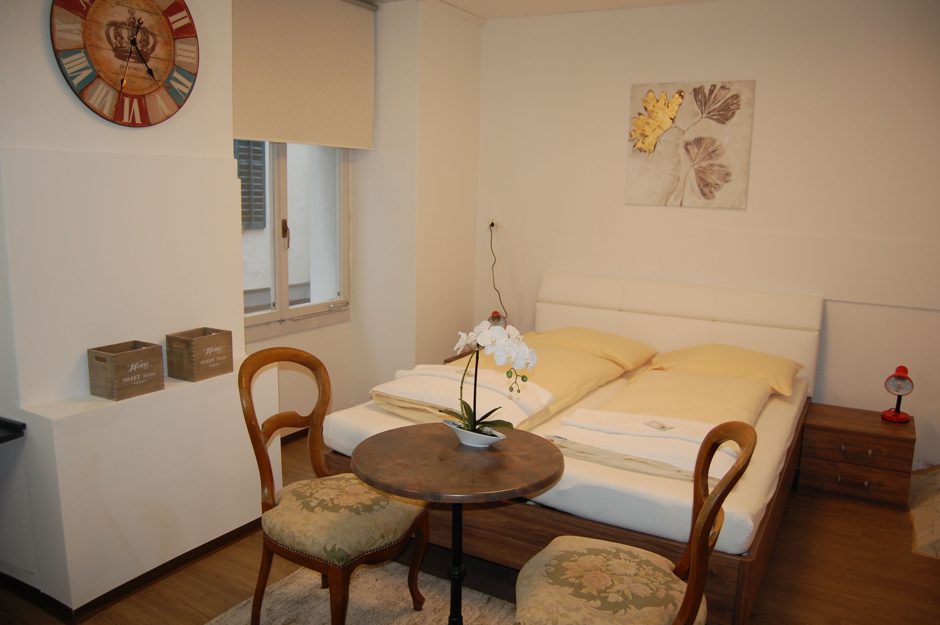 Une chambre à coucher avec un lit, deux chaises, une petite table, une horloge et une fenêtre.