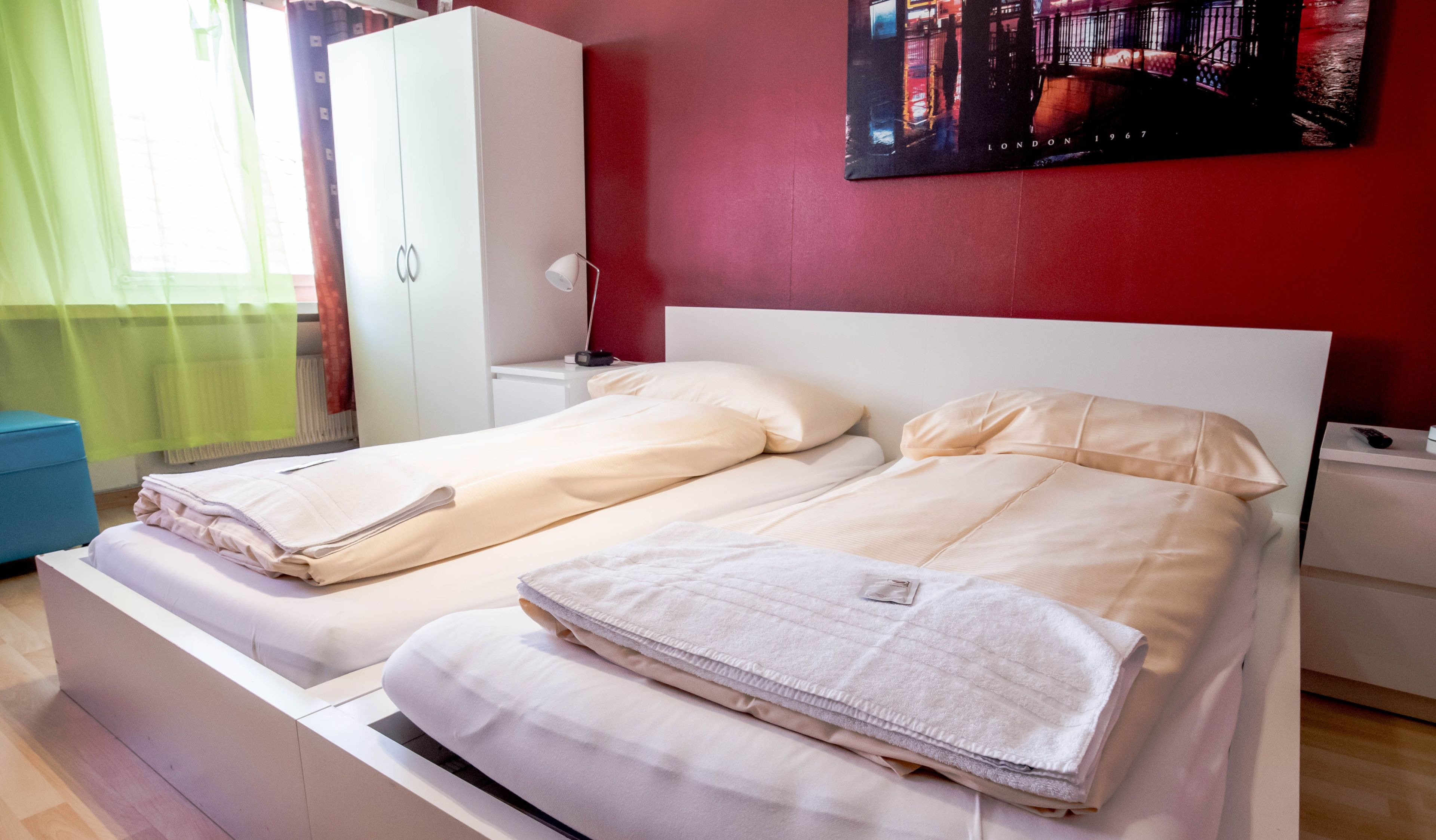 A double bed with white bedding, a nightstand with a lamp, and a red wall with a cityscape painting.