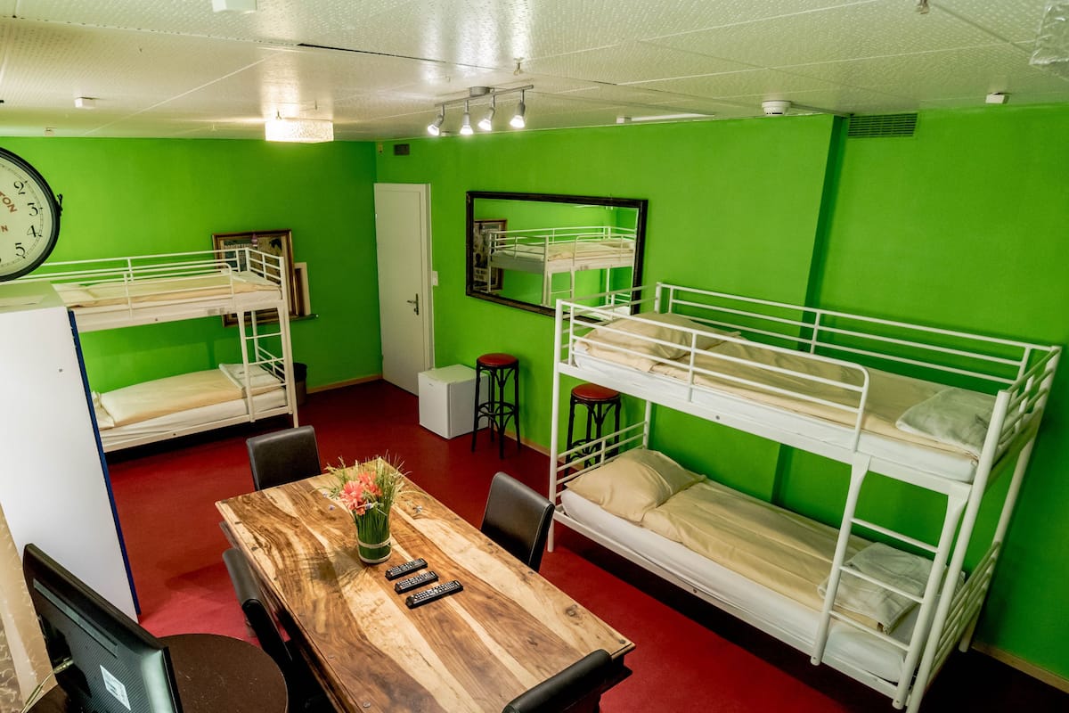 A dormitory room with bunk beds, a wooden table with a vase of flowers, and a clock on the wall.