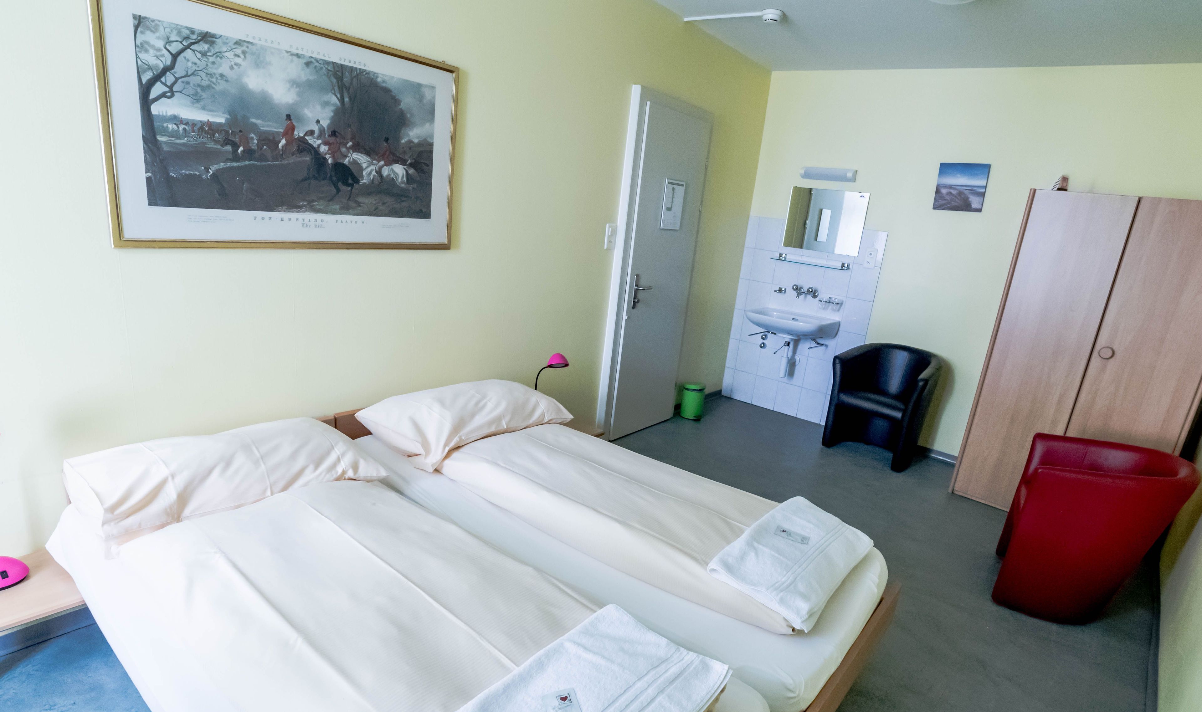 Ein Hotelzimmer mit zwei Betten, einem Badezimmer mit Waschbecken und Spiegel sowie einem gerahmten historischen Gemälde an der Wand.
