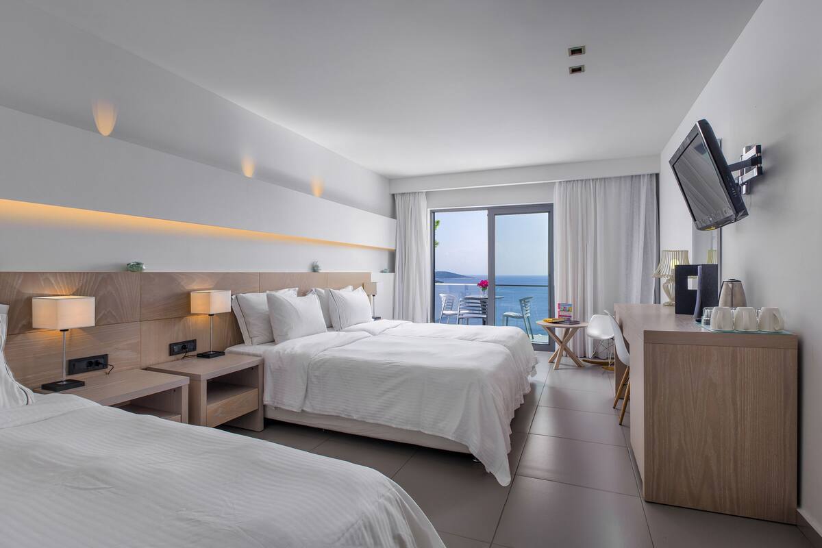 A modern hotel room with two beds, a desk, a TV, and a view of the sea.