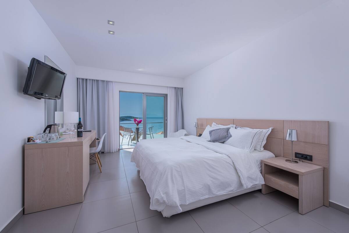 A modern hotel room with a large bed, a desk with a flat-screen TV, a bedside table with a lamp, and a view of the sea through the window.