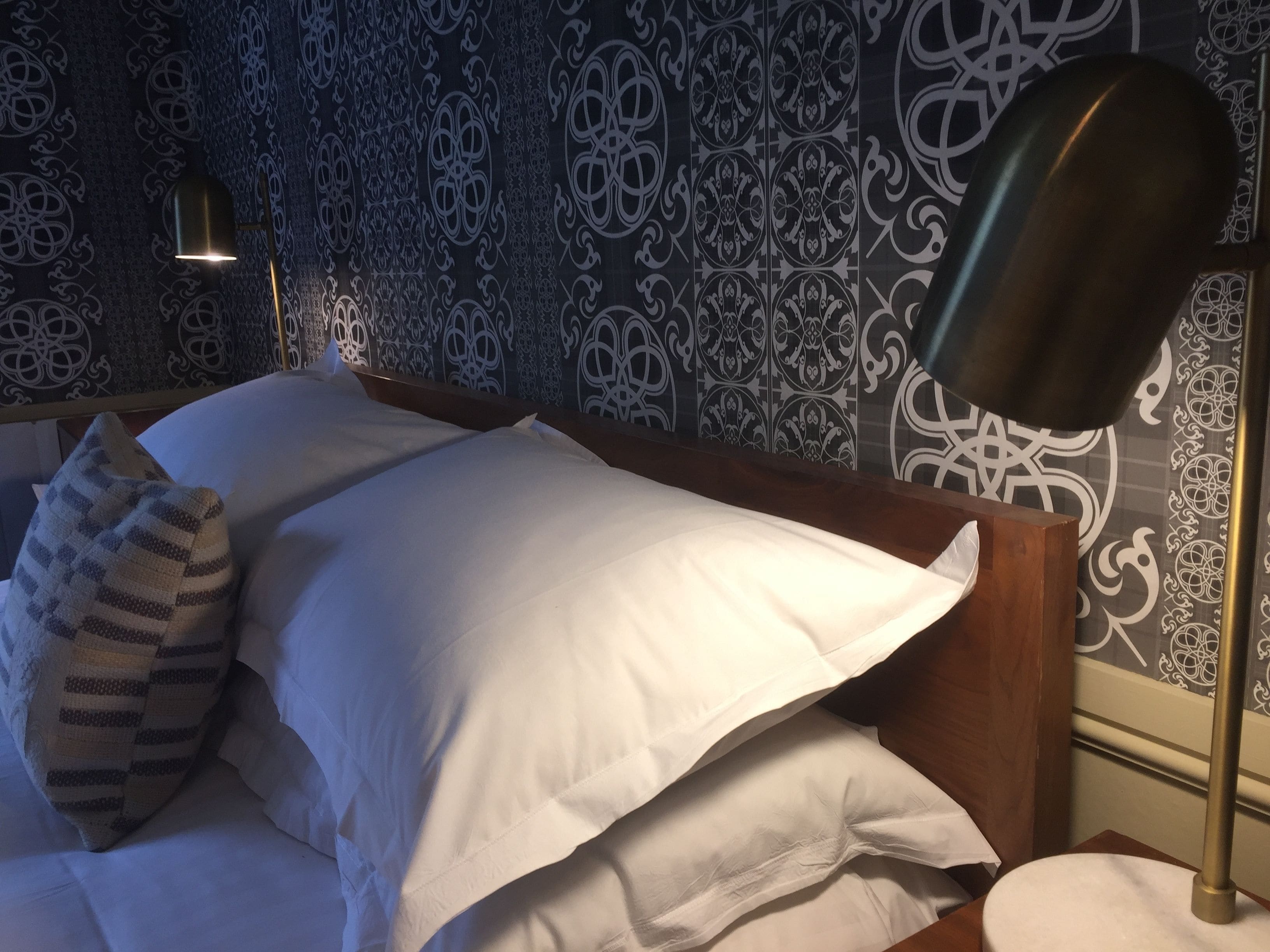 A neatly made bed with white linens, a striped pillow, and a modern floor lamp.