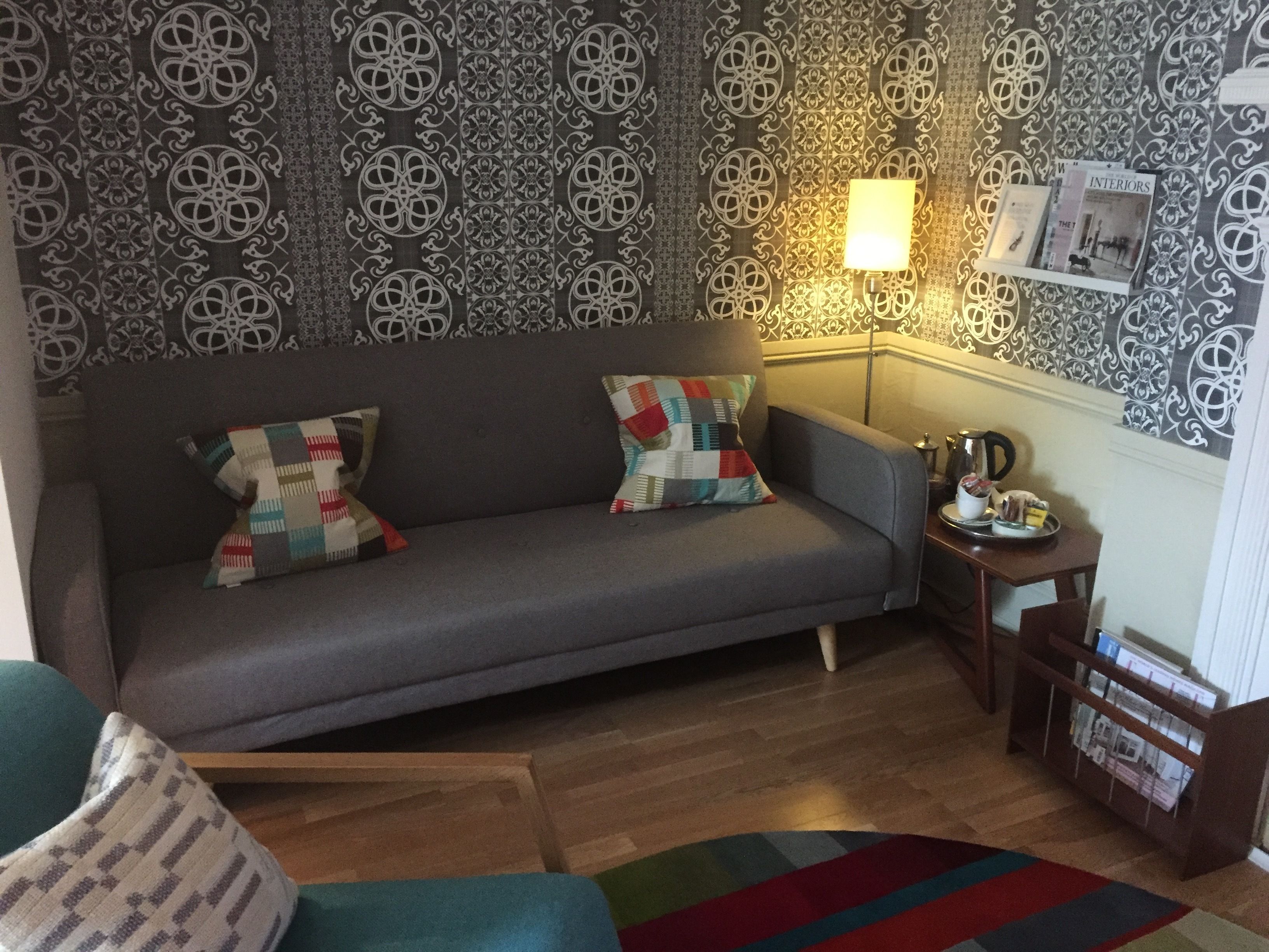 A living room with a grey sofa, two patterned cushions, a small side table with a lamp, a coffee pot, and a magazine rack.