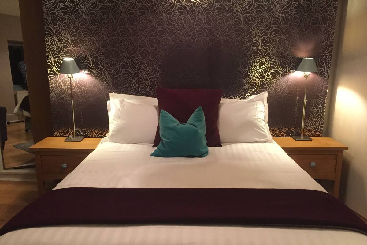 A neatly made bed with white and teal pillows, a dark red accent pillow, and a maroon throw blanket. There are two bedside tables with lamps, and a patterned wallpaper backdrop.