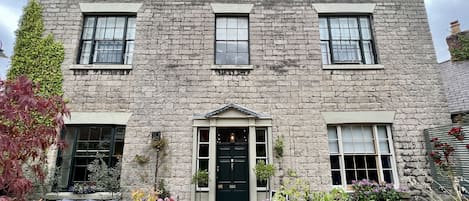 Un edificio de piedra con entrada central, ventanas grandes y plantas en macetas alrededor de la entrada.