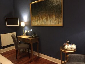 A hotel room with a wooden desk, a chair, a bedside table with a lamp, a painting on the wall, and a radiator.