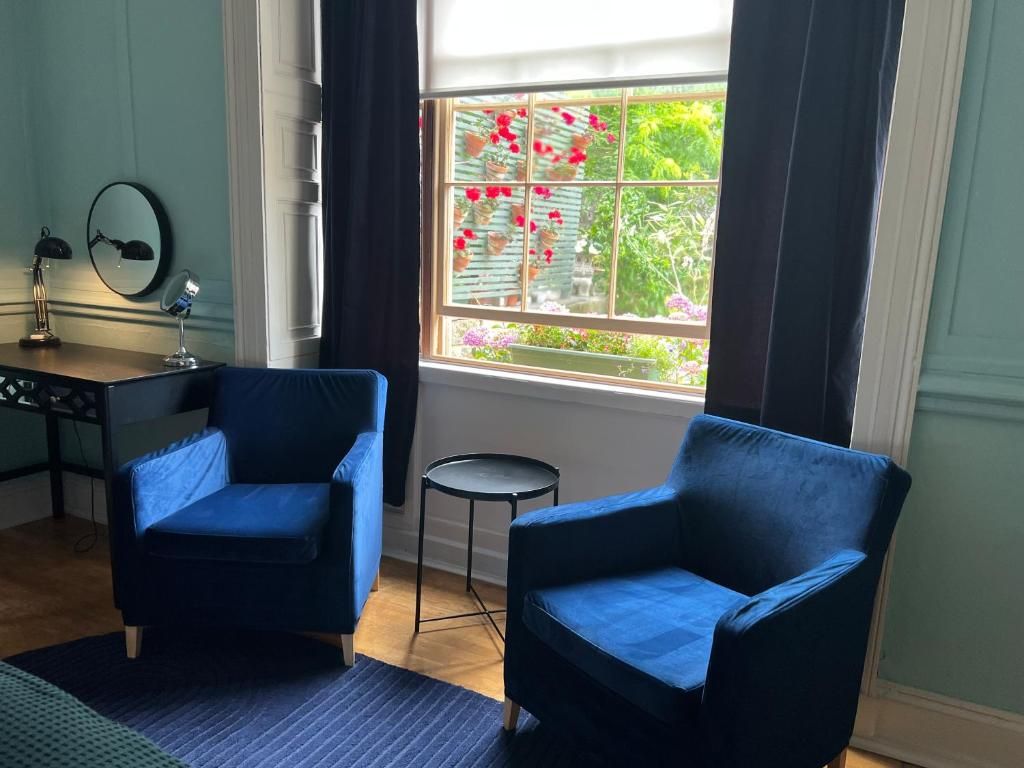 A room with two blue armchairs, a small round table, a desk with a lamp, and a window with a view of greenery and flowers.