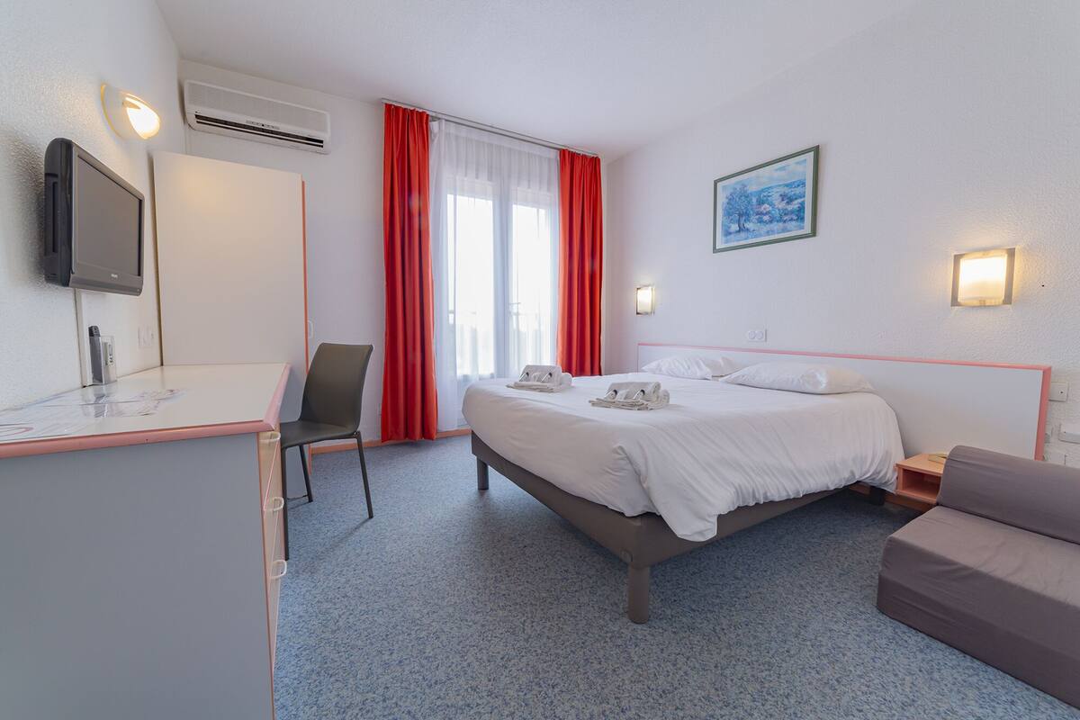 Ein Hotelzimmer mit Bett, Schreibtisch, Sessel, Sofa, einem Gemälde und einem Fenster mit roten Vorhängen.