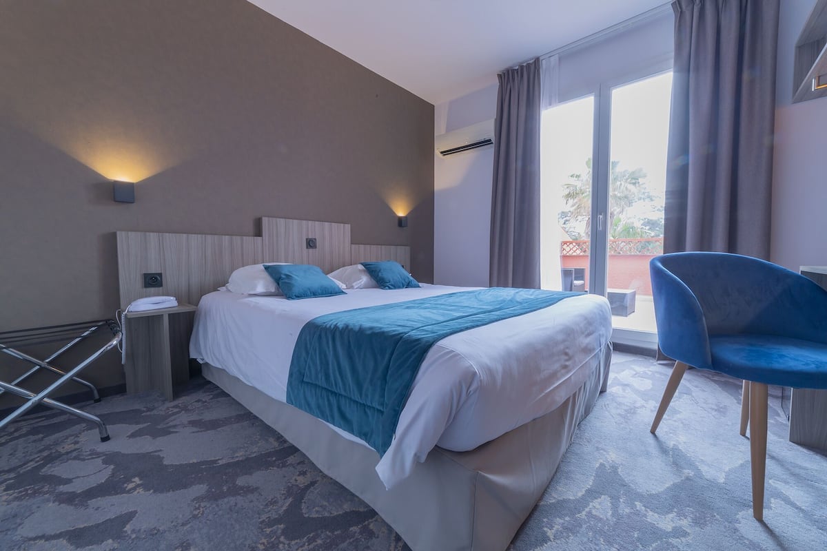 Ein Hotelzimmer mit Bett, Sessel, Nachttisch, Klimaanlage und Blick ins Freie.