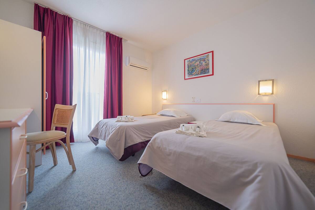Ein Hotelzimmer mit zwei Betten, einem Schreibtisch, einem Stuhl und einem Vorhangfenster.