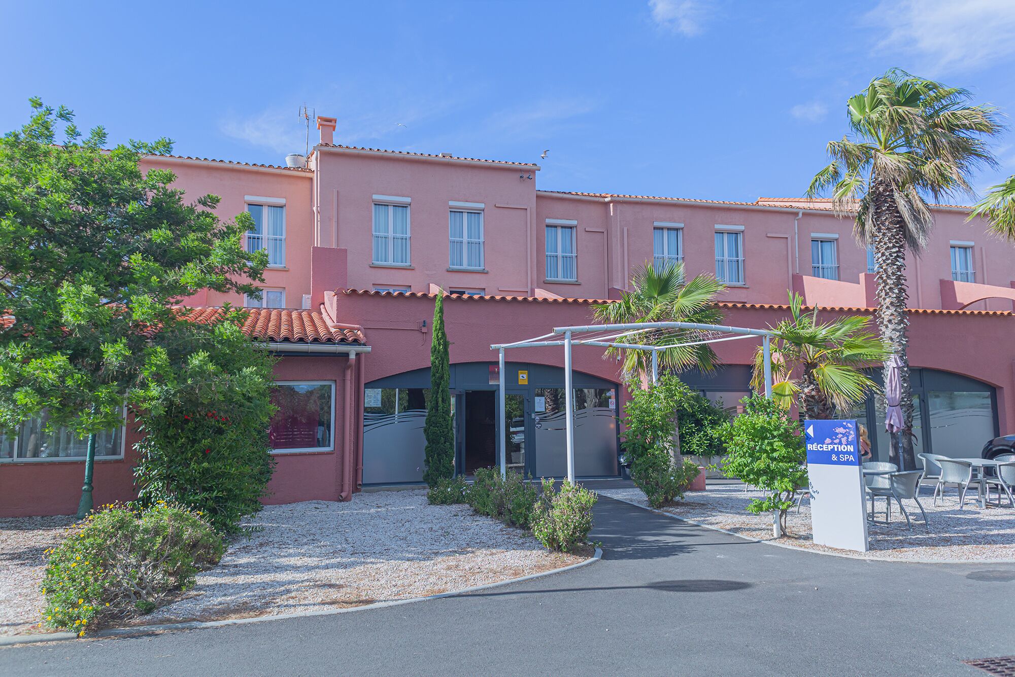 A building with a reception and spa area, surrounded by greenery and palm trees.