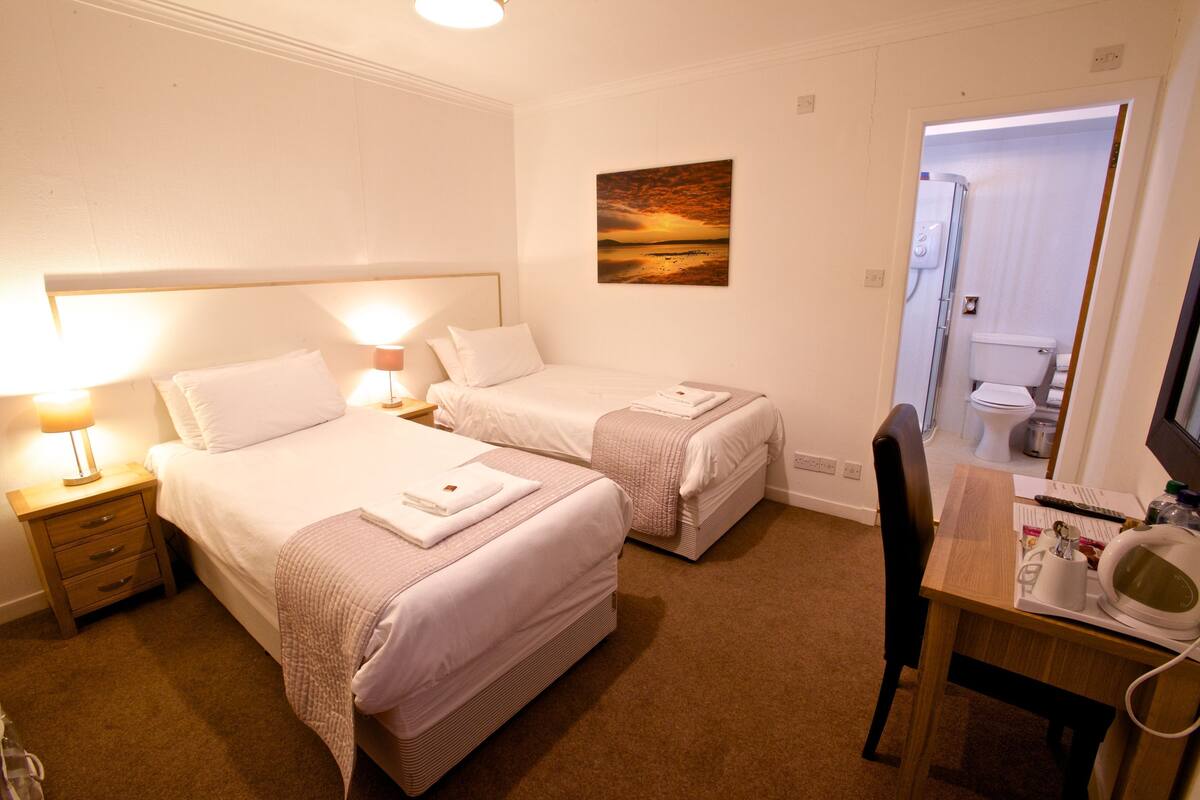 A hotel room with two beds, a desk with a chair, a painting on the wall, and a bathroom visible through an open door.