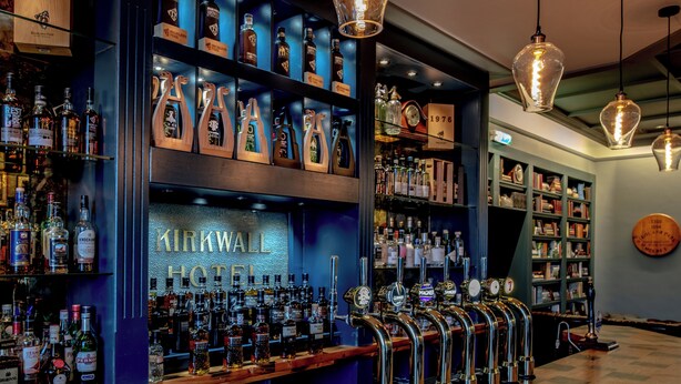 Eine Bar mit zahlreichen Spirituosenflaschen, einem Schild mit der Aufschrift "Kirkwall Hotel" und mehreren Bierhähnen.