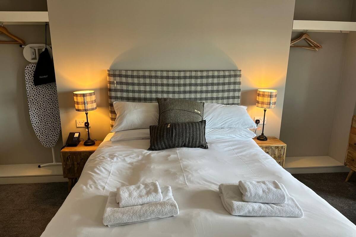 A neatly made bed with white linens and two folded towels, flanked by two bedside tables with lamps and a phone.