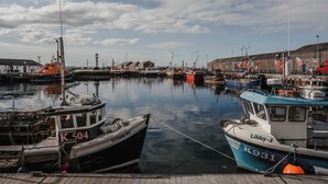 Ein Hafen mit vertäuten Booten am Kai, ein Leuchtturm im Hintergrund und Gebäude entlang der Uferlinie.
